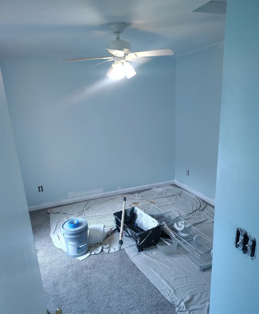 A room with light blue walls, a white ceiling fan, and paint supplies on a drop cloth over gray carpet.